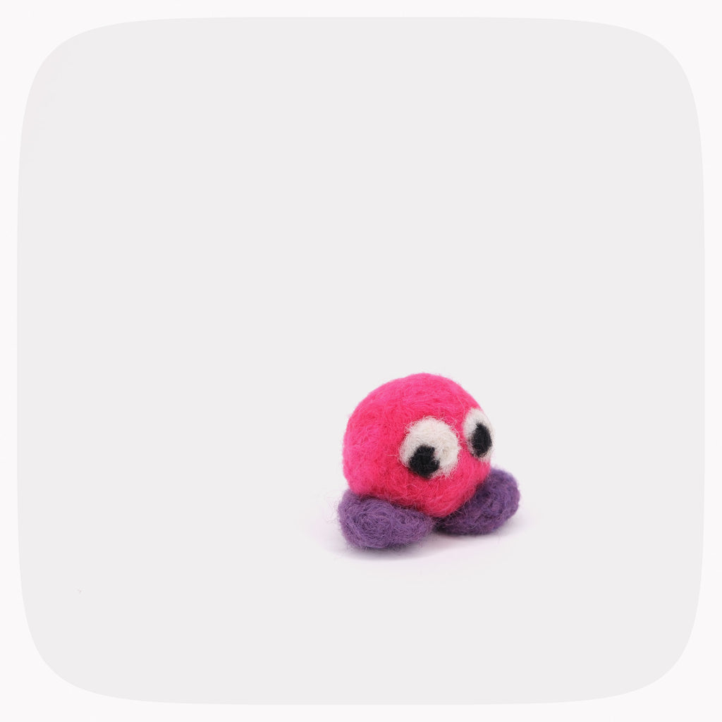 A small felted pink ball with purple feet and eyes looking left.