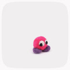 A small felted pink ball with purple feet and eyes looking left.