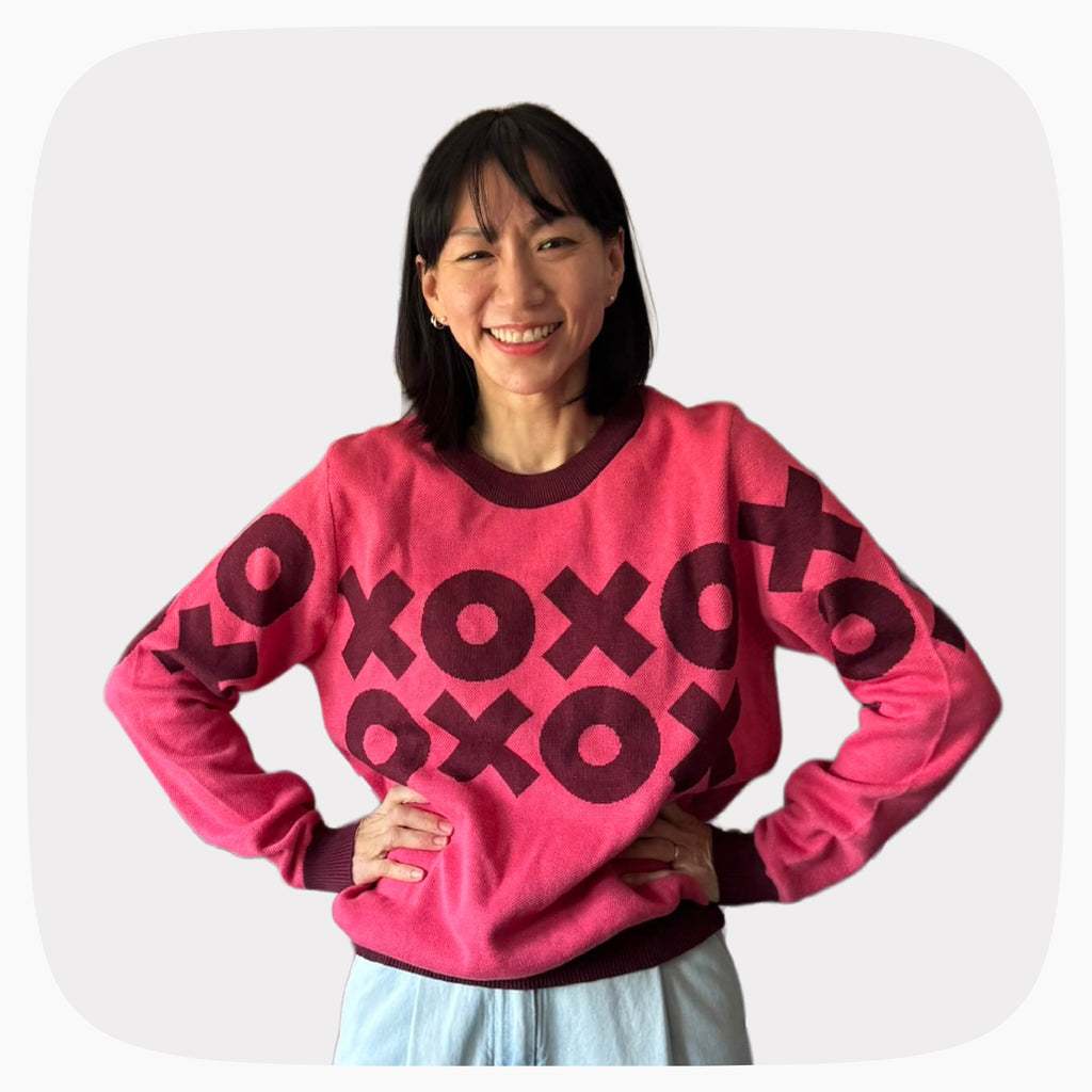 A woman wearing a pink graphic knit sweater with a dark red XOXO pattern across the chest.