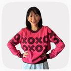 A woman wearing a pink graphic knit sweater with a dark red XOXO pattern across the chest.