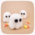 Small felted white ghost figurines with black eyes photographed on a kraft brown background with candy corn in the background.