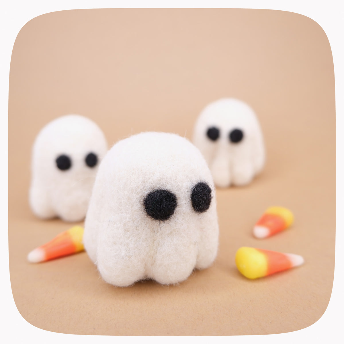 Small felted white ghost figurines with black eyes photographed on a kraft brown background with candy corn in the background.